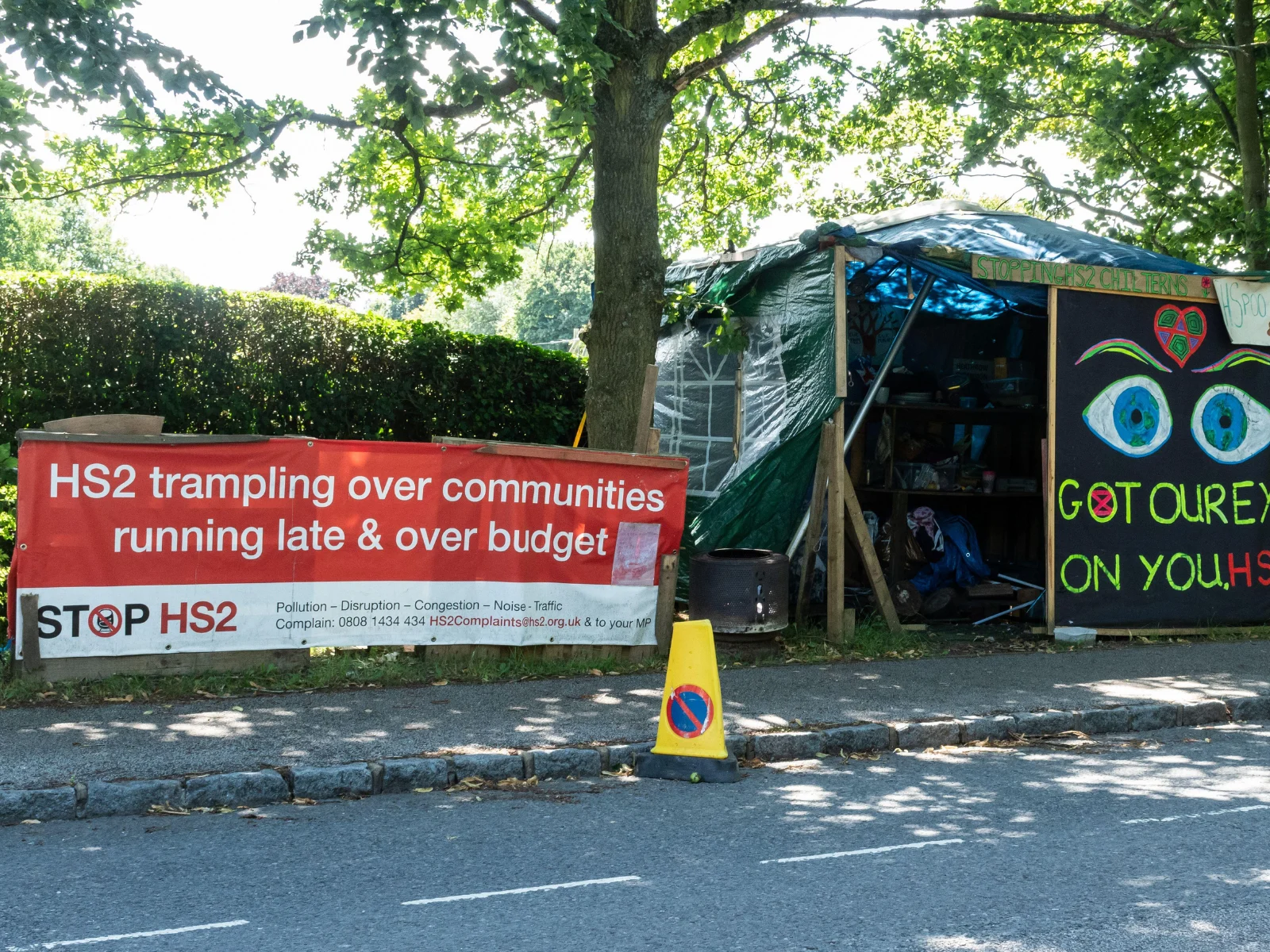 Roadside protest camp in the Chilterns with a red banner reading "HS2 trampling over communities, running late and over budget."